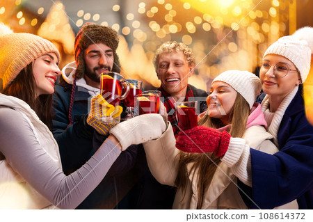 Group portrait of adorable smiling people in cozy fair atmosphere standing outdoor and clinking cup of hot warming drink. Group portrait of adorable smiling people in cozy fair atmosphere standing outdoor and clinking cup of hot warming drink. 108614372