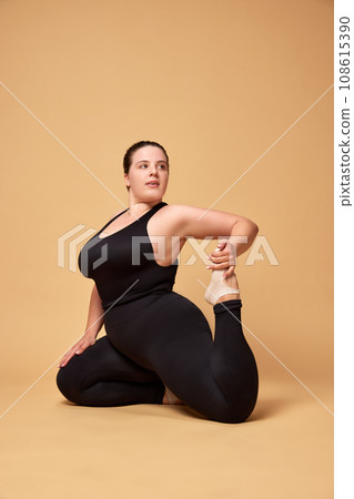 Young overweight woman in black sportswear training, sitting and stretching against beige studio background. Sport, body-positivity Young overweight woman in black sportswear training, sitting and stretching against beige studio background. Sport, body-positivity 108615390