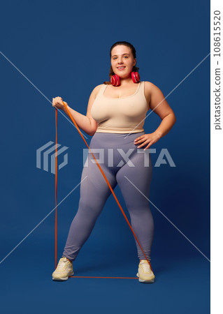 Fat, overweigh young woman training with elastic bands, fitness expanders against blue studio background. Sport, body-positivity Fat, overweigh young woman training with elastic bands, fitness expanders against blue studio background. Sport, body-positivity 108615520