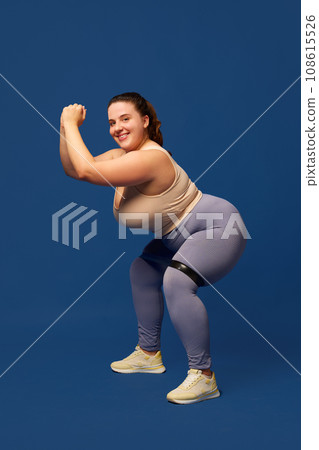 Young overweigh woman training with fitness elastic band, doing squats against blue studio background. Sport, body-positivity 108615526