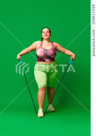 Young overweigh woman in sportswear training, doing exercises with fitness expanders against green studio background. Sport, body-positivity 108615599