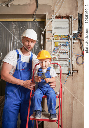 Little boy in safety helmet sitting on ladder and holding construction tool while man standing beside kid. Male worker and child posing near electrical switchboard in apartment under renovation. 108616041