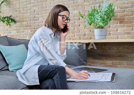 Young female university student studying at home, talking on cell phone Young female university student studying at home, talking on cell phone 108616272