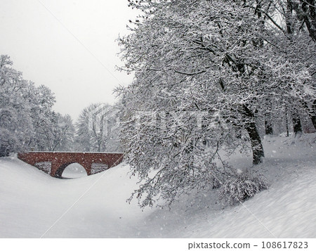 A small brick bridge over a snow-covered moat in the Zamosc city park. Snow-covered trees, snowfall. Winter landscape. A small brick bridge over a snow-covered moat in the Zamosc city park. Snow-covered trees, snowfall. Winter landscape. 108617823