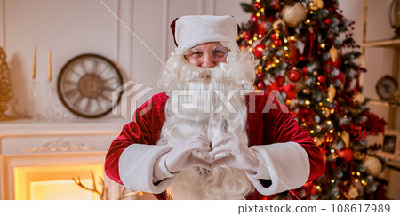 Santa Claus forming shape of heart with fingers near fireplace and christmas tree with gifts Santa Claus forming shape of heart with fingers near fireplace and christmas tree with gifts 108617989