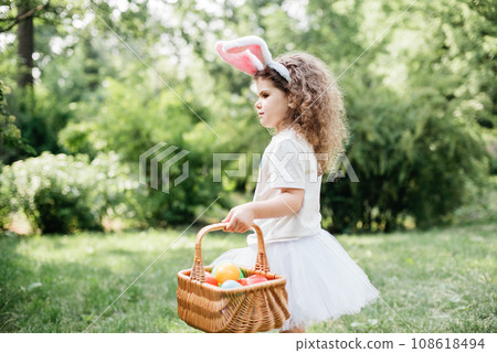 Easter egg hunt. Girl child Wearing Bunny Ears Running To Pick Up Egg In Garden. Easter tradition. Baby with basket full of colorful eggs. Easter egg hunt. Girl child Wearing Bunny Ears Running To Pick Up Egg In Garden. Easter tradition. Baby with basket full of colorful eggs. 108618494