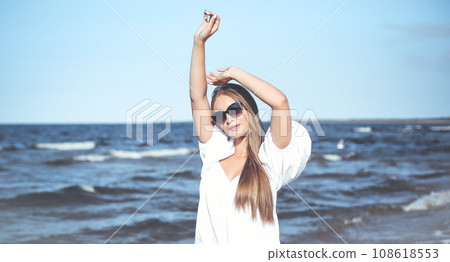 Happy blonde woman is on the ocean beach in a white dress and sunglasses, raising hands 108618553
