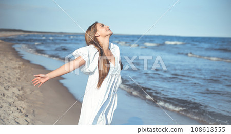 Happy smiling beautiful woman is on the ocean beach in a white summer dress, open arms 108618555