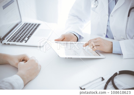 Doctor and patient sitting opposite each other at the desk in clinic. The focus is on female physician's hands using tablet computer, close up. Medicine concept Doctor and patient sitting opposite each other at the desk in clinic. The focus is on female physician's hands using tablet computer, close up. Medicine concept 108618623