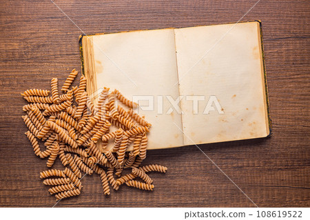 Open blank recipe book and raw whole grain fusilli pasta on wooden table. Top view. Open blank recipe book and raw whole grain fusilli pasta on wooden table. Top view. 108619522