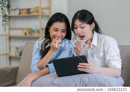 Two young asian women using tablet while sitting together at home. 108622223