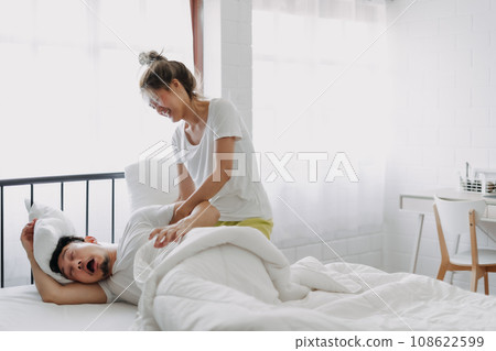 Funny couple wife try to wake lazy husband up in white bedroom. Funny couple wife try to wake lazy husband up in white bedroom. 108622599