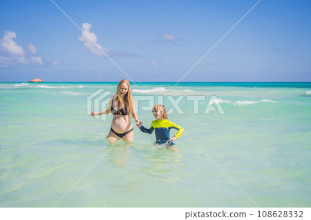 A radiant pregnant mother and her excited son share a tender moment on a serene, snow-white beach, celebrating family love amidst nature's beauty A radiant pregnant mother and her excited son share a tender moment on a serene, snow-white beach, celebrating family love amidst nature's beauty 108628332