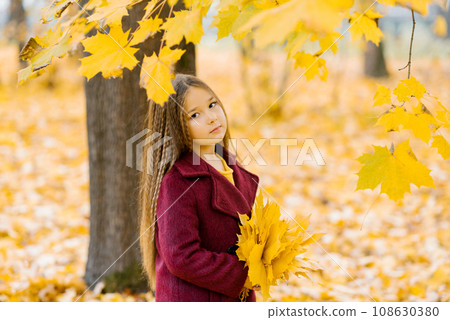 Autumn street portrait of a beautiful happy child girl walking in a park or forest in a coat, holding a bouquet of yellow leaves in her hands 108630380