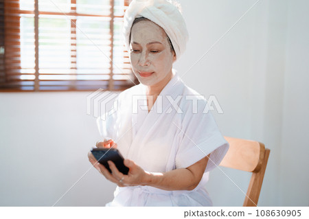 Portrait of elderly woman doing face spa treatment and using mobile phone 108630905