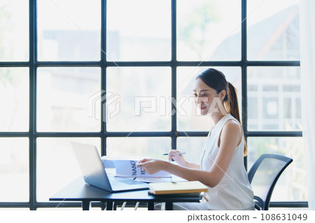 Portrait of a thoughtful Asian businesswoman looking at financial statements and making marketing plans using a computer on her desk. 108631049