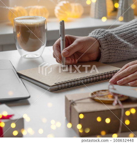 Young woman writing christmas cards, makes order on laptop. Female picking gifts online purchases at cozy home among gift boxes and packages. Winter sales, Black Friday. Christmas discount promotions 108632448