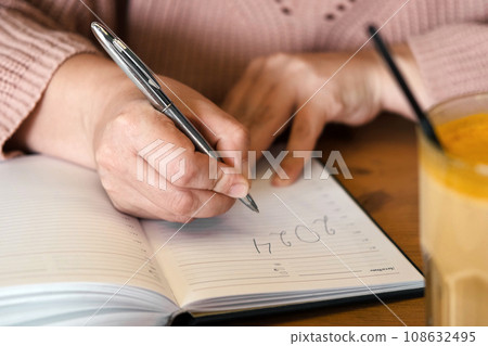 Woman's hand writing 2024. New Year Routine, resolutions on desk. Cafe, restaurant interior background. 2024 goals list with notebook, coffee cup on wooden table. Action, checklist, idea concept. Woman's hand writing 2024. New Year Routine, resolutions on desk. Cafe, restaurant interior background. 2024 goals list with notebook, coffee cup on wooden table. Action, checklist, idea concept. 108632495