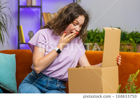 Happy young woman shopper unpacking cardboard box delivery parcel online shopping purchase at home Happy young woman shopper unpacking cardboard box delivery parcel online shopping purchase at home 108633750