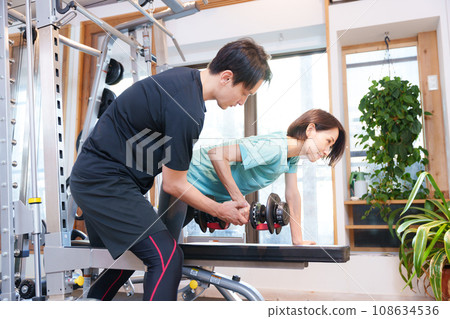 A woman and trainer (dumbbells) exercising at a sports gym A woman and trainer (dumbbells) exercising at a sports gym 108634536