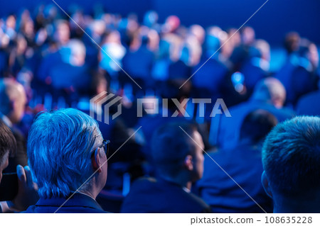 Businessmen listening to speaker giving presentation in illuminated auditorium at export forum Businessmen listening to speaker giving presentation in illuminated auditorium at export forum 108635228