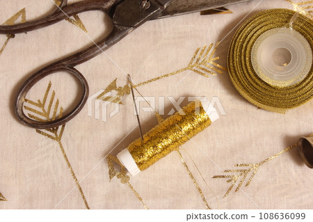 Spool of Gold Thread and Scissors With Thimble on Metallic Fabric 108636099