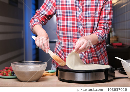 Woman cooking delicious crepe on electric pancake maker in kitchen, closeup Woman cooking delicious crepe on electric pancake maker in kitchen, closeup 108637026