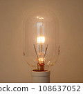 An oblong incandescent light bulb with a filament that glows slightly and gently. 108637052