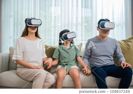 Happy family have mother father and daughter watching movie or playing video game, game entertainment innovation technology, Asian family wear vr glasses headsets excited sitting sofa in living room 108637289