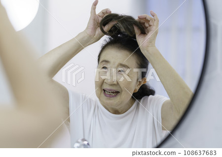 Smiling senior woman putting on a partial wig in front of the mirror 108637683