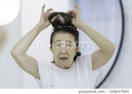 A senior woman with a troubled face putting on a partial wig in front of the mirror A senior woman with a troubled face putting on a partial wig in front of the mirror 108637684