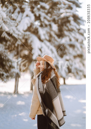 Smiling woman enjoying winter moments in snowy park. Young woman wearing hat, plaid scarf and coat. 108638335