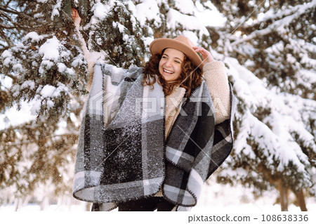 Smiling woman enjoying winter moments in snowy park. Young woman wearing hat, plaid scarf and coat. Smiling woman enjoying winter moments in snowy park. Young woman wearing hat, plaid scarf and coat. 108638336