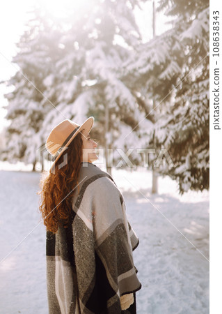 Smiling woman enjoying winter moments in snowy park. Young woman wearing hat, plaid scarf and coat. 108638343