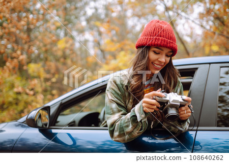 Beautiful woman takes pictures on camera from the car window. Smiling woman enjoying autumn weather. 108640262