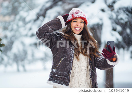 Happy woman in a red hat with a phone takes a selfie, blogs in the winter forest. Concept of nature. 108641049