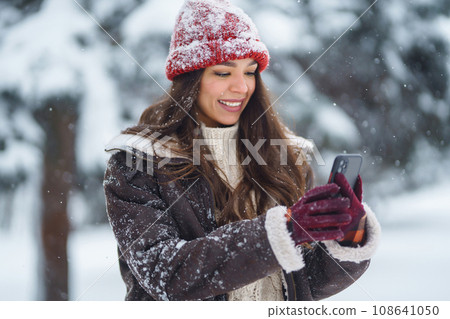 Happy woman in a red hat with a phone takes a selfie, blogs in the winter forest. Concept of nature. 108641050