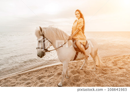 A white horse and a woman in a dress stand on a beach, with the 108641983