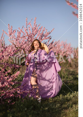 Woman blooming peach orchard. Against the backdrop of a picturesque peach orchard, a woman in a long pink dress and hat enjoys a peaceful walk in the park, surrounded by the beauty of nature. 108642014