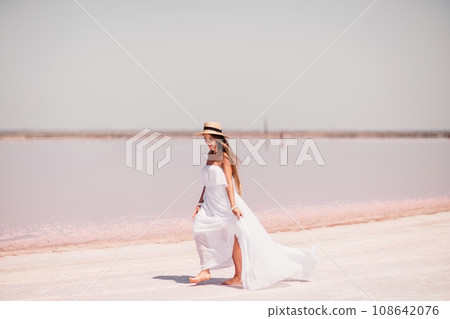 Woman in pink salt lake. She in a white dress and hat enjoys the scenic view of a pink salt lake as she walks along the white, salty shore, creating a lasting memory. 108642076
