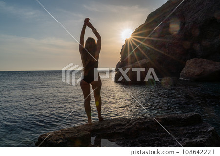 Woman tourist enjoying the sunset over the sea mountain landscape. Sits outdoors on a rock above the sea. She is wearing jeans and a blue hoodie. Healthy lifestyle, harmony and meditation Woman tourist enjoying the sunset over the sea mountain landscape. Sits outdoors on a rock above the sea. She is wearing jeans and a blue hoodie. Healthy lifestyle, harmony and meditation 108642221
