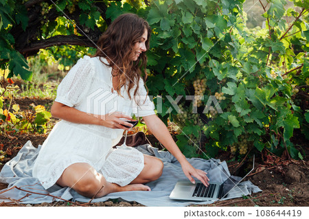 A woman under a vineyard with a laptop and wine in a glass 108644419