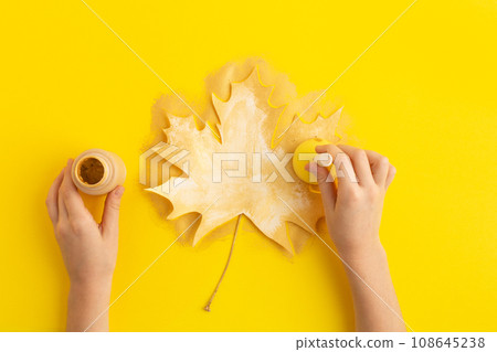 Child hand paints autumn leaves in gold, children occupation. Autumn concept flatlay on yellow. DIY 108645238