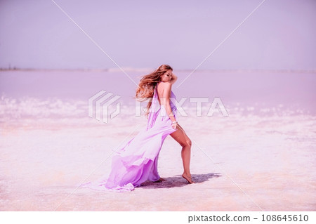 Woman pink salt lake. Against the backdrop of a pink salt lake, a woman in a long pink dress takes a leisurely stroll along the white, salty shore, capturing a wanderlust moment. 108645610