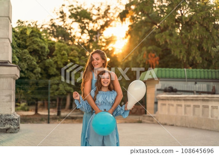 Mother daughter family sunset. Portrait of mother and daughter in blue dresses with flowing long hair against the backdrop of sunset. A woman hugs and presses the girl to her, holding balloons in her 108645696
