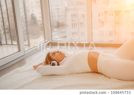 Side view portrait of relaxed woman listening to music with headphones lying on carpet at home. She is dressed in a white tracksuit. 108645733