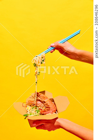 Unrecognizable woman eating delicious hot WOK with shrimps with chopstick from paper box isolated yellow studio background Unrecognizable woman eating delicious hot WOK with shrimps with chopstick from paper box isolated yellow studio background 108646296
