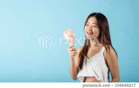 Asian beauty young woman holding pink portable electric mini fan near her face studio shot isolated on blue background, Female hand hold small plastic fan handheld she enjoying cool wind blowing 108646817