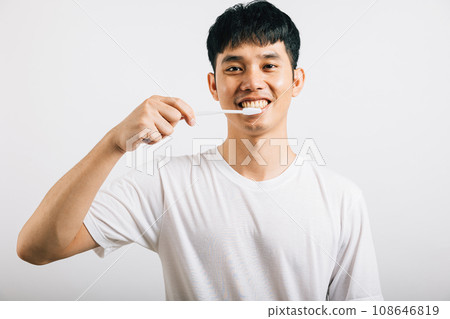 An Asian young man confidently brushes his teeth in the morning, promoting dental health. Portrait of a happy Thai teen with a toothbrush in a studio shot isolated on a white background. 108646819