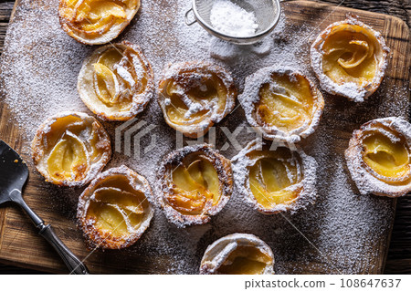 Mini tarts made of puff pastry and sliced apples sprinkled with powdered sugar on wooden cutting board 108647637